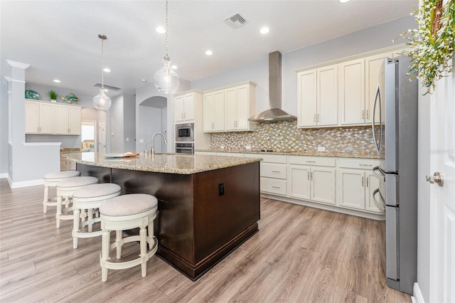 kitchen with appliances with stainless steel finishes, an island with sink, wall chimney exhaust hood, pendant lighting, and light hardwood / wood-style flooring