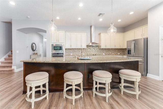 kitchen featuring a center island with sink, wall chimney range hood, stainless steel appliances, and light stone counters