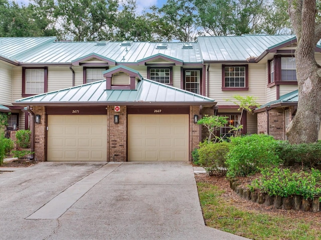 townhome / multi-family property featuring driveway, a standing seam roof, an attached garage, brick siding, and metal roof