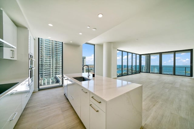 kitchen with expansive windows, sink, a kitchen island with sink, white cabinetry, and a water view