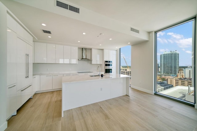 kitchen with double oven, light hardwood / wood-style flooring, wall chimney range hood, and white cabinetry