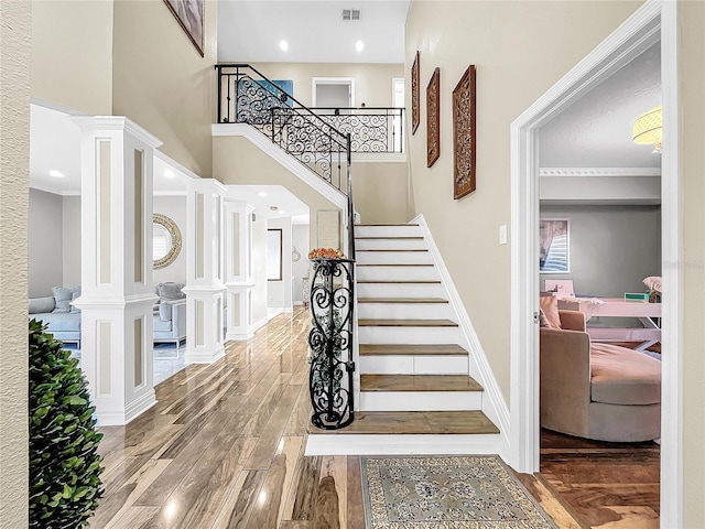 stairs featuring wood-type flooring, crown molding, a high ceiling, and ornate columns