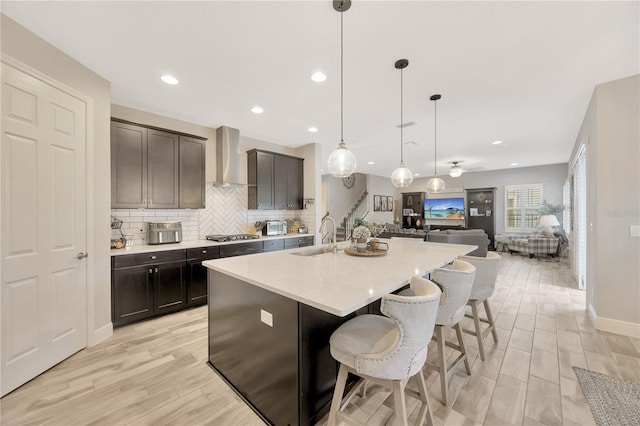 kitchen featuring wall chimney range hood, sink, hanging light fixtures, light hardwood / wood-style flooring, and an island with sink