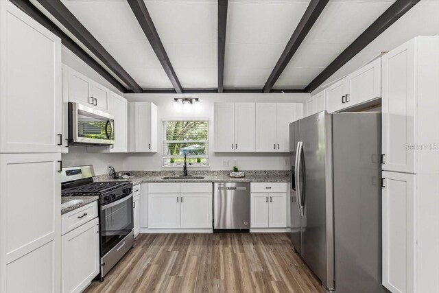kitchen featuring light wood finished floors, stainless steel appliances, white cabinets, a sink, and beamed ceiling