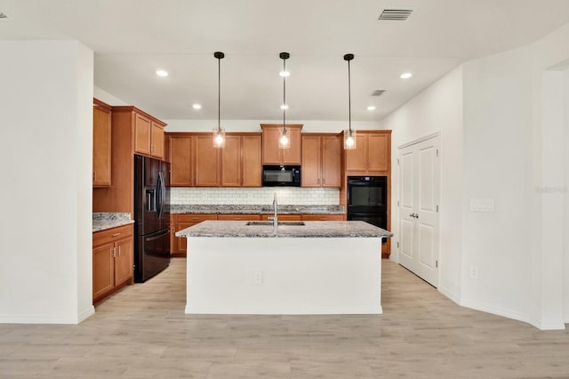 kitchen with light wood-type flooring, light stone counters, a kitchen island with sink, black appliances, and decorative light fixtures