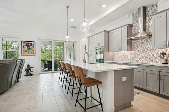 kitchen featuring pendant lighting, stainless steel double oven, a kitchen island with sink, and wall chimney range hood