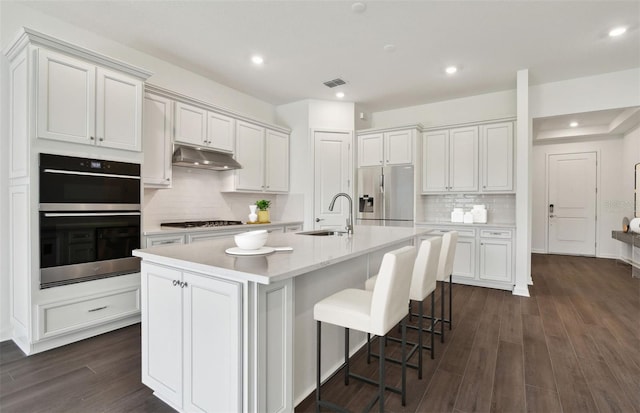 kitchen featuring sink, stainless steel refrigerator with ice dispenser, double oven, a kitchen island with sink, and white cabinets