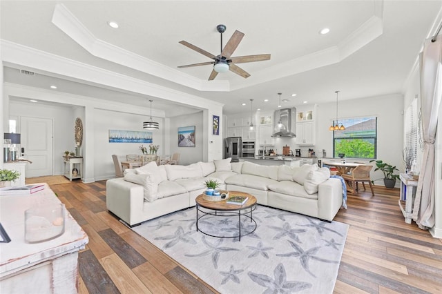 living room featuring wood-type flooring, ornamental molding, ceiling fan with notable chandelier, and a raised ceiling