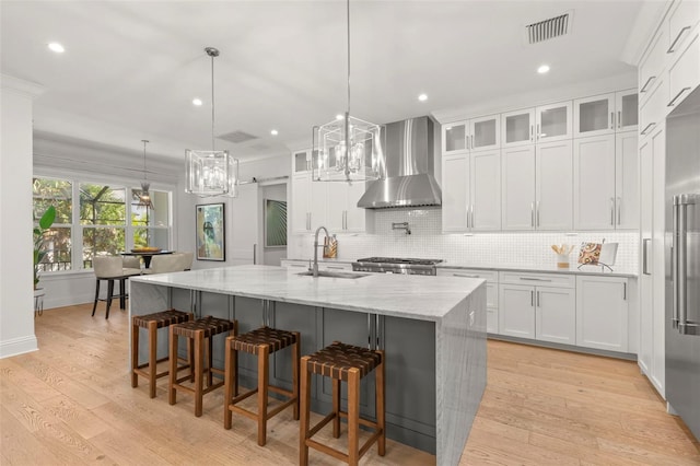 kitchen featuring a large island with sink, white cabinets, wall chimney exhaust hood, light wood-type flooring, and decorative light fixtures