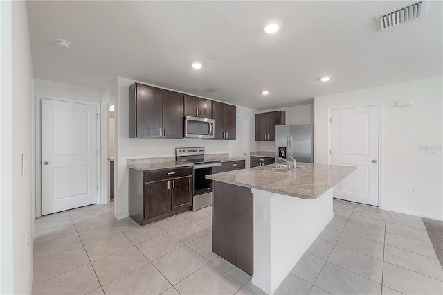 kitchen featuring dark brown cabinetry, light tile patterned floors, a textured ceiling, a center island with sink, and appliances with stainless steel finishes