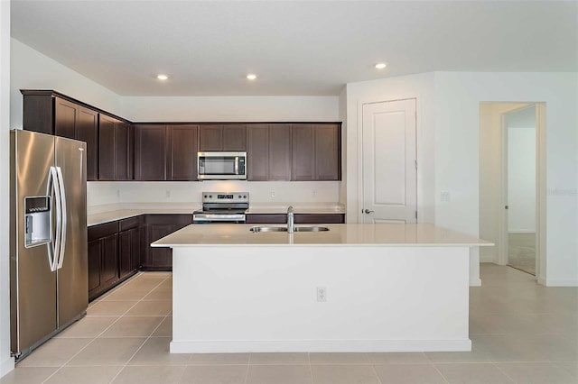 kitchen with appliances with stainless steel finishes, an island with sink, sink, light tile patterned floors, and dark brown cabinetry