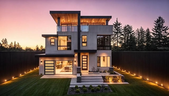 back of property at dusk featuring a yard, fence, a balcony, and stucco siding