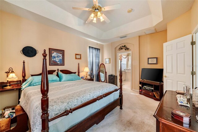 bedroom with ceiling fan, a raised ceiling, light carpet, and ensuite bathroom