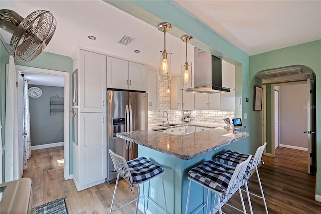 kitchen with stainless steel fridge, white cabinetry, stone countertops, kitchen peninsula, and wall chimney exhaust hood