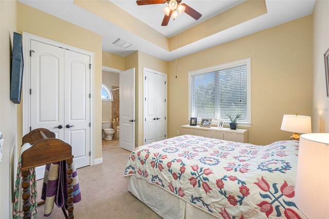 bedroom featuring a raised ceiling, ensuite bath, ceiling fan, and light colored carpet