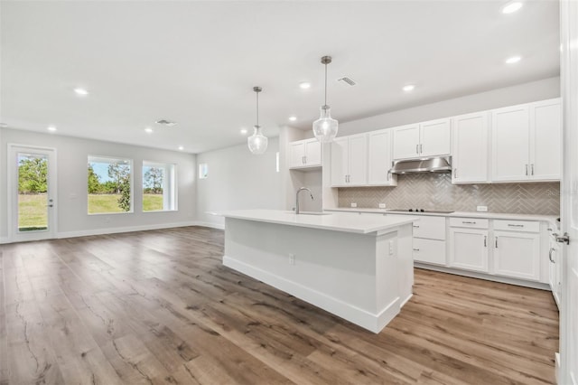 kitchen with black electric stovetop, a kitchen island with sink, sink, white cabinets, and hanging light fixtures