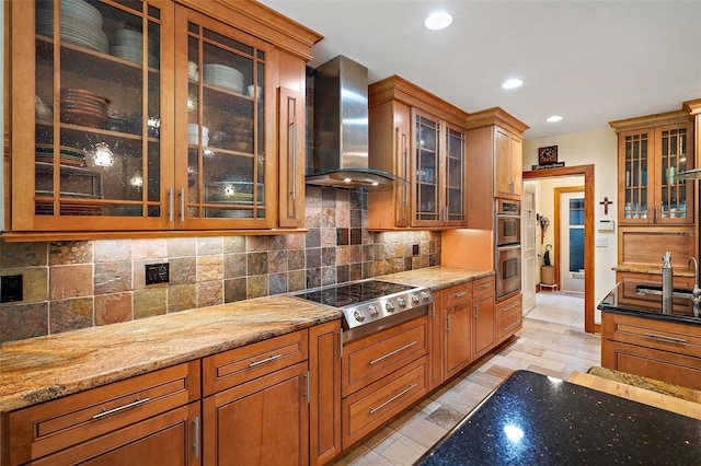 kitchen featuring light wood-type flooring, light stone counters, tasteful backsplash, wall chimney range hood, and appliances with stainless steel finishes