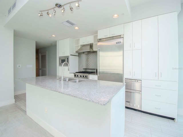 kitchen featuring light stone counters, white cabinets, built in appliances, ventilation hood, and a kitchen island with sink