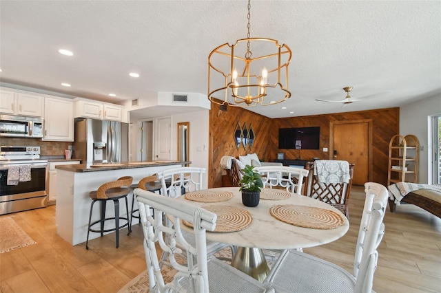 dining space with a textured ceiling, ceiling fan with notable chandelier, wood walls, and light hardwood / wood-style floors