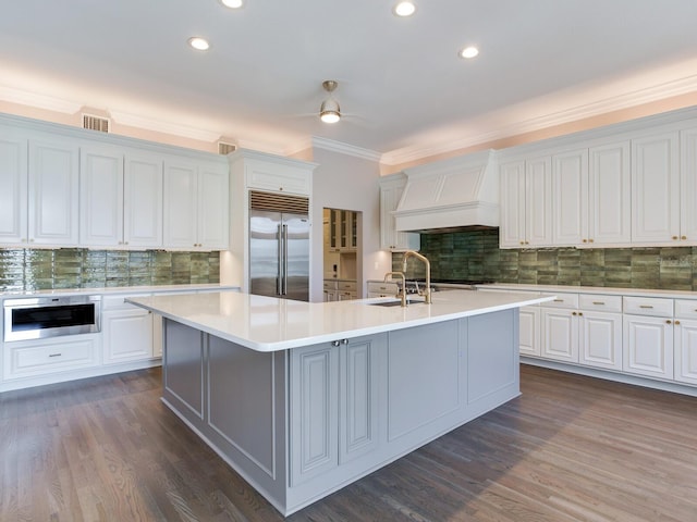 kitchen featuring a kitchen island with sink, dark wood-type flooring, appliances with stainless steel finishes, white cabinetry, and premium range hood