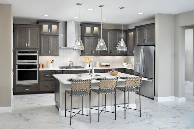 kitchen featuring wall chimney exhaust hood, a center island with sink, appliances with stainless steel finishes, and decorative light fixtures