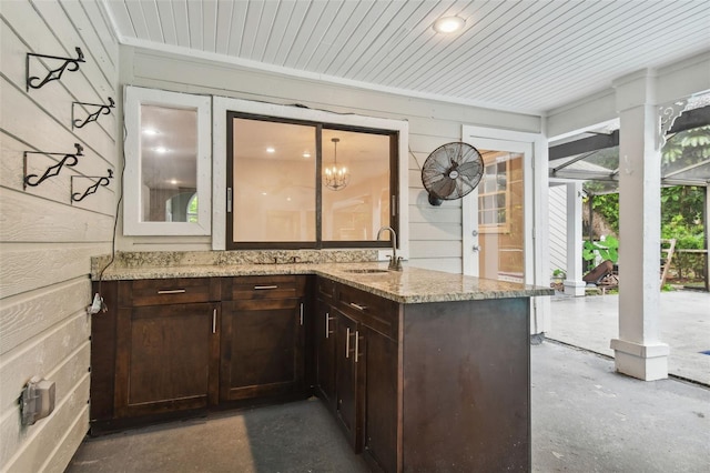 bar with light stone counters, sink, wooden walls, and dark brown cabinetry