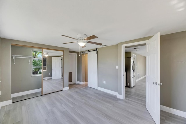 unfurnished bedroom featuring a closet, light hardwood / wood-style floors, a barn door, ceiling fan, and stainless steel fridge