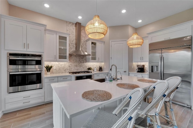 kitchen featuring an island with sink, white cabinets, hanging light fixtures, wall chimney exhaust hood, and appliances with stainless steel finishes