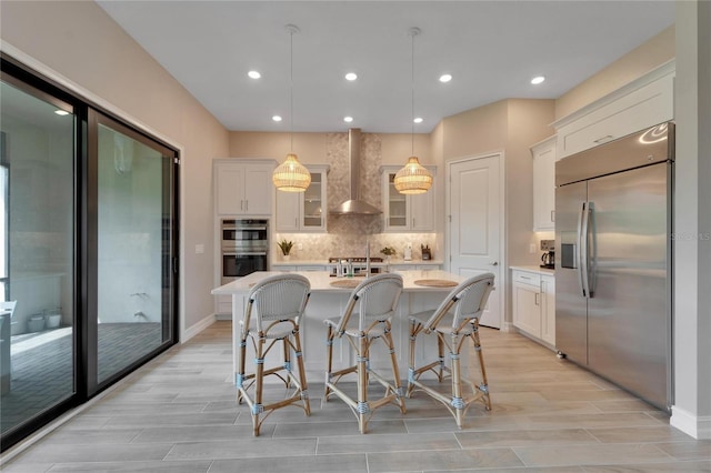 kitchen with white cabinets, decorative light fixtures, a center island with sink, wall chimney exhaust hood, and stainless steel appliances