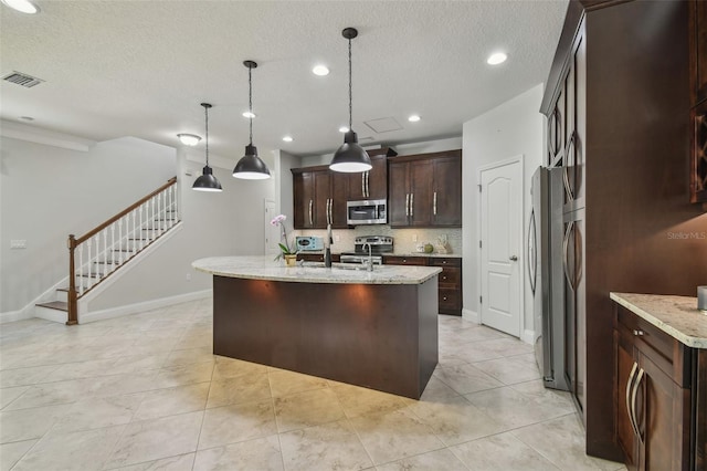 kitchen featuring pendant lighting, a textured ceiling, a kitchen island with sink, appliances with stainless steel finishes, and light stone countertops