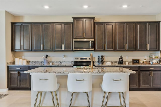 kitchen with an island with sink, stainless steel appliances, dark brown cabinets, and light stone countertops