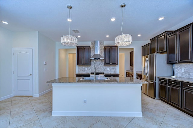 kitchen with an island with sink, stainless steel fridge, decorative light fixtures, wall chimney exhaust hood, and decorative backsplash