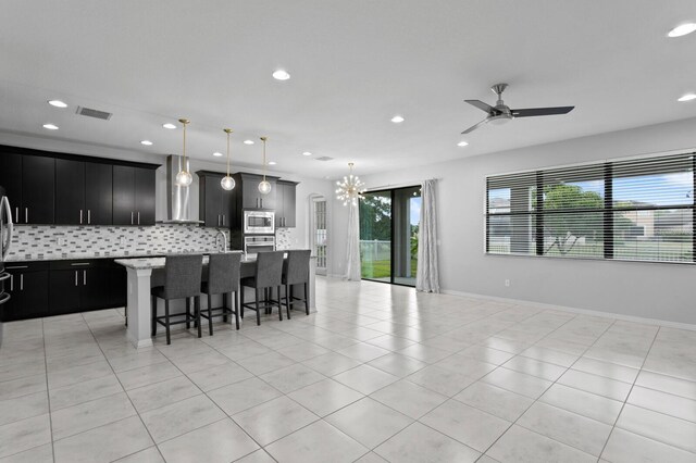 kitchen featuring stainless steel appliances, wall chimney range hood, backsplash, an island with sink, and decorative light fixtures