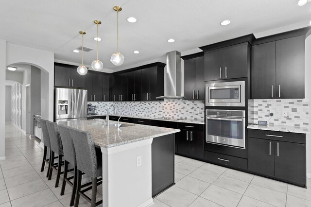 kitchen featuring appliances with stainless steel finishes, sink, wall chimney range hood, pendant lighting, and an island with sink