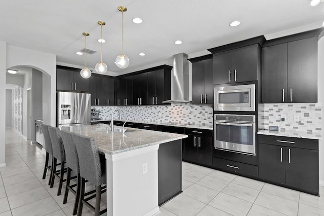 kitchen featuring wall chimney exhaust hood, stainless steel appliances, a sink, decorative light fixtures, and dark cabinets