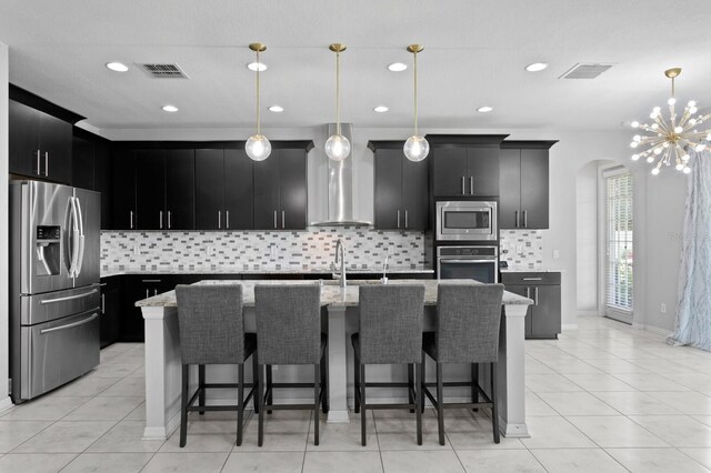 kitchen featuring pendant lighting, wall chimney exhaust hood, stainless steel appliances, and a kitchen island with sink