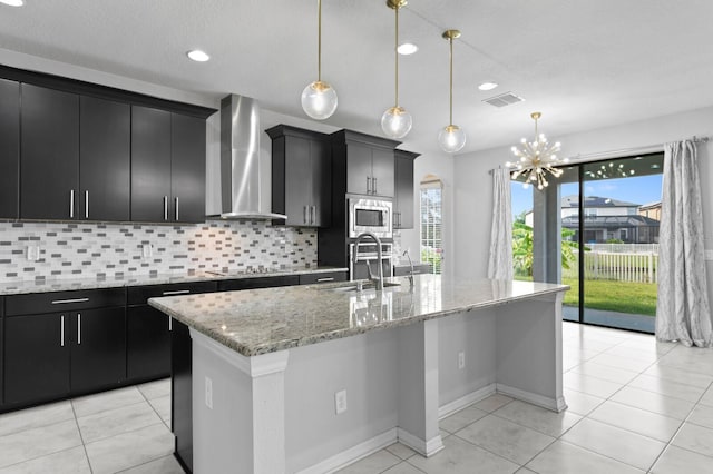 kitchen with a kitchen island with sink, stainless steel microwave, a sink, dark cabinets, and wall chimney range hood
