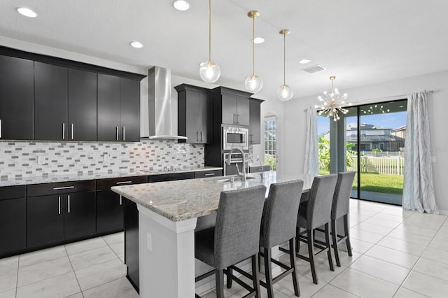 kitchen with a sink, stainless steel microwave, wall chimney range hood, visible vents, and dark cabinets