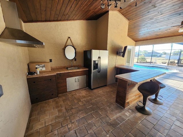 kitchen featuring stainless steel fridge, rail lighting, wood ceiling, sink, and wall chimney range hood