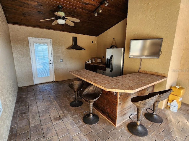 kitchen with tile counters, wooden ceiling, stainless steel fridge with ice dispenser, track lighting, and a kitchen bar