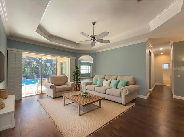 living room featuring a tray ceiling, dark wood-style flooring, baseboards, arched walkways, and a ceiling fan