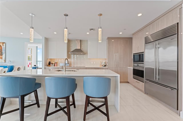 kitchen featuring a kitchen island with sink, built in appliances, sink, wall chimney exhaust hood, and decorative light fixtures