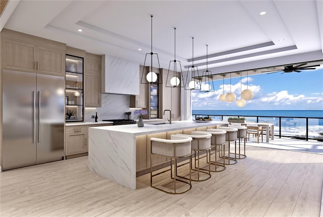 kitchen with light hardwood / wood-style floors, a kitchen island with sink, stainless steel built in fridge, a water view, and a raised ceiling