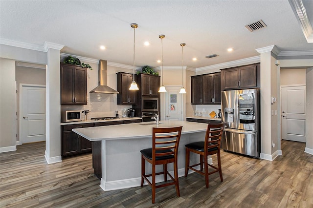 kitchen with an island with sink, wall chimney exhaust hood, hanging light fixtures, stainless steel appliances, and light countertops