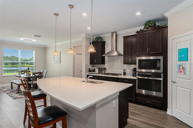 kitchen featuring stainless steel appliances, sink, wall chimney range hood, a center island with sink, and hanging light fixtures