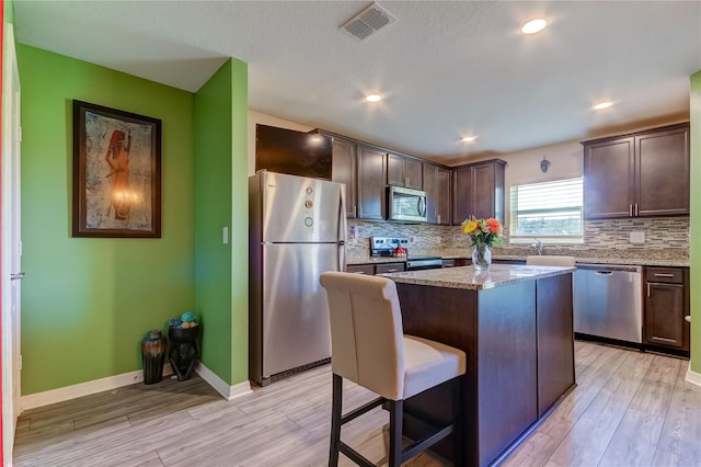 kitchen featuring appliances with stainless steel finishes, a kitchen island, light stone countertops, light wood-type flooring, and dark brown cabinets