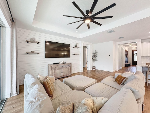 living room with a tray ceiling, light wood finished floors, visible vents, and ceiling fan