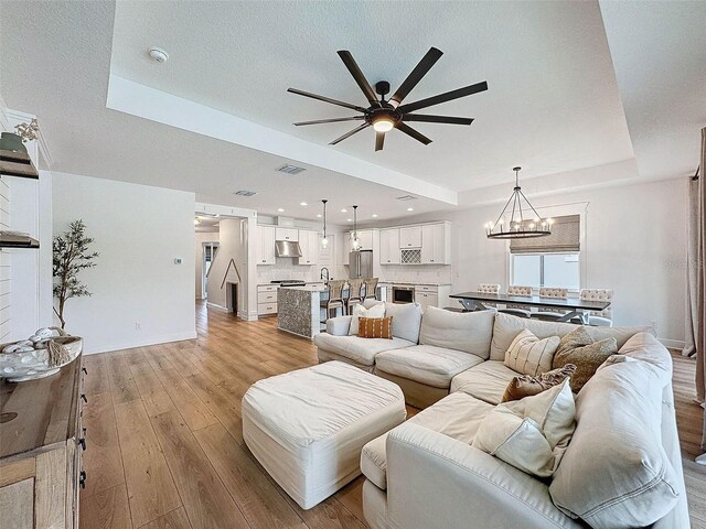 living room with visible vents, baseboards, light wood-type flooring, ceiling fan with notable chandelier, and a raised ceiling