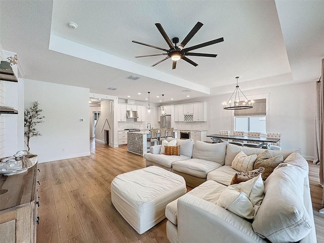 living room featuring ceiling fan with notable chandelier, baseboards, a raised ceiling, visible vents, and light wood-style flooring