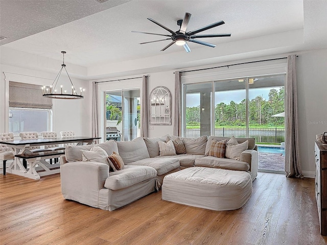 living room featuring a tray ceiling, light wood-style flooring, a healthy amount of sunlight, and ceiling fan with notable chandelier